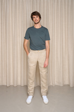 Modèle homme portant le jeans en chanvre L'Aurelien en chanvre naturel, coupe droite avec poches italiennes, couleur naturelle, fermeture éclair recyclée et bouton en bois de buis, tissé en Auvergne Rhône-Alpes et assemblé près de Lyon, chaussé de baskets blanches, sur fond uni et neutre