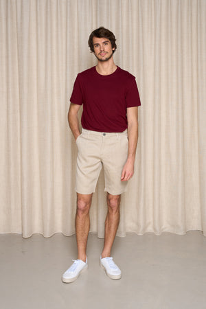 Homme portant le bermuda Cabellio en lin de couleur lin naturel avec coupe confortable sur les hanches et longueur jusqu'au genou, accompagné d'un t-shirt bordeaux et de baskets blanches, sur fond de rideau beige - Le Gaulois Jeans