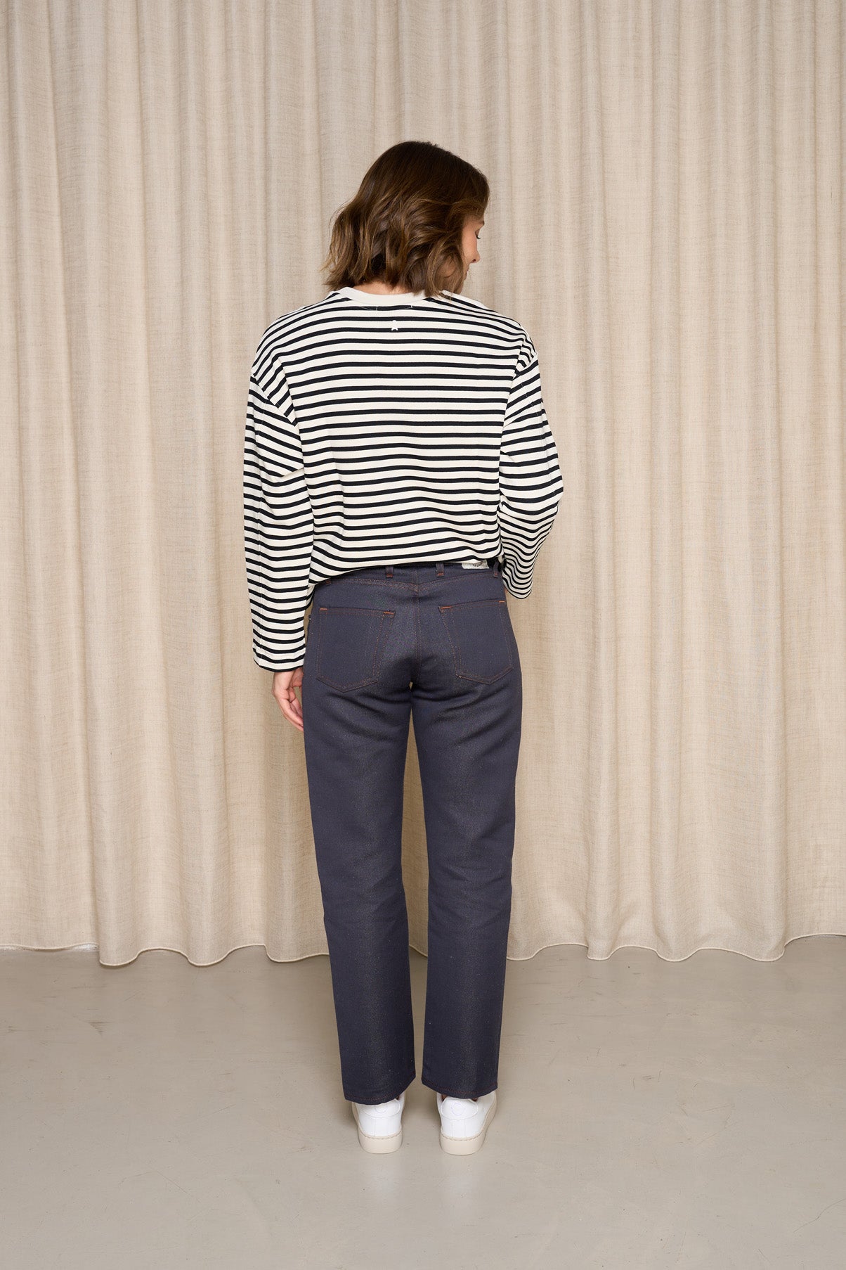 Femme de dos portant le jeans en chanvre et coton bio Isla pour femme à coupe droite, couleur bleu avec fermeture à glissière et liseré rouge visible sur le revers, associé à un haut à rayures et chaussures blanches sur fond de rideau clair - Le Gaulois jeans