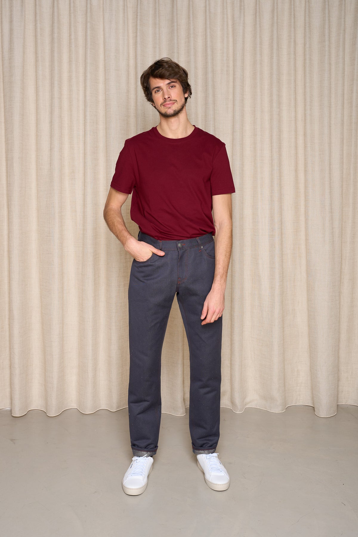 Homme présentant le jeans en chanvre et coton bio Isla pour homme couleur bleu à coupe droite, taille haute avec fermeture à glissière et liseré rouge intérieur, porté avec un t-shirt bordeaux et des baskets blanches, sur fond de rideau beige - Le Gaulois jeans