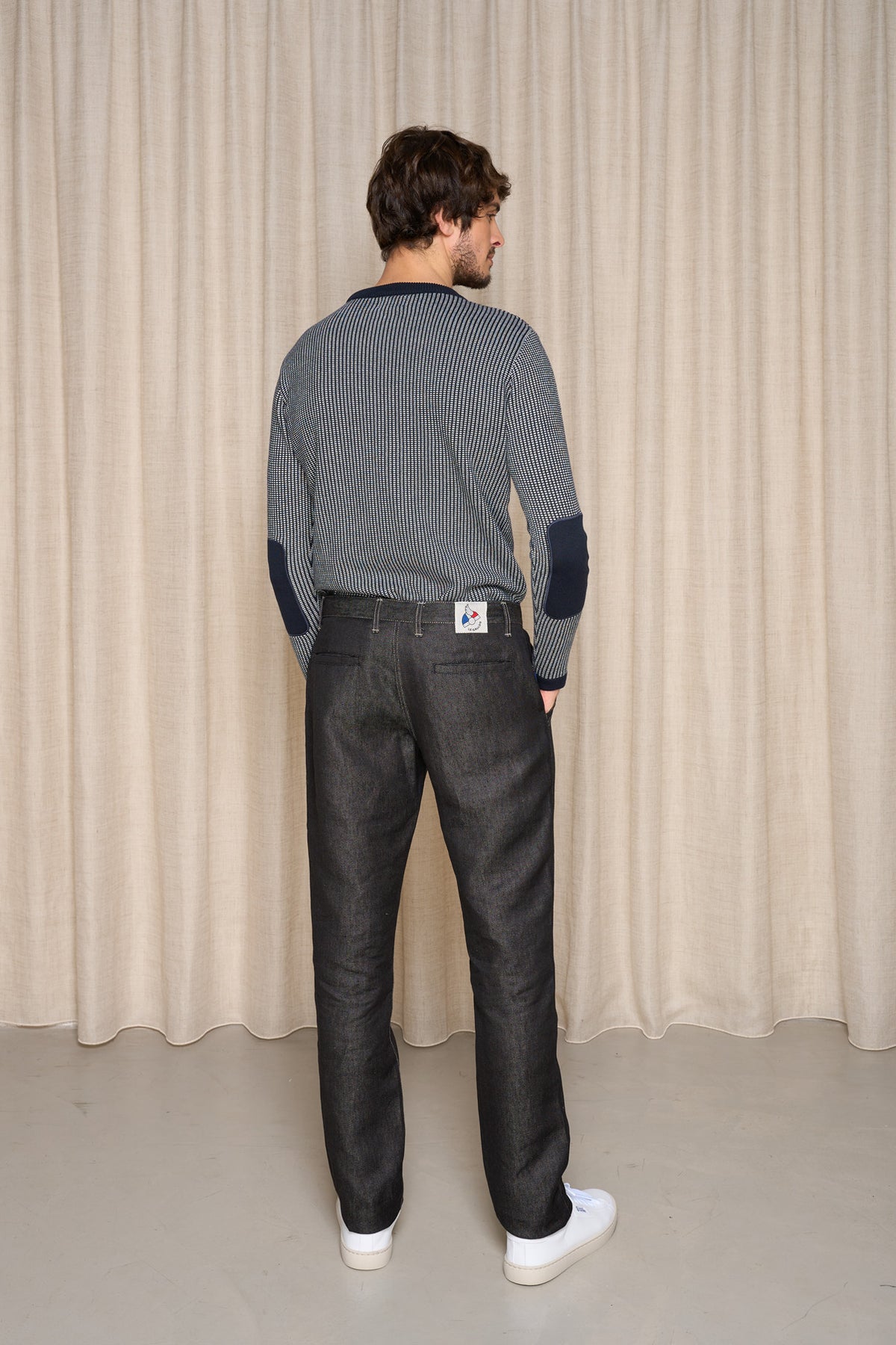 Homme de dos portant le jeans en lin Lugdunum Noir coupe chino, taille haute et legèrement effilé aux chevilles, accompagné d'un pull bleu à motif géométrique et des baskets blanches, sur fond de rideau beige - Le Gaulois jeans