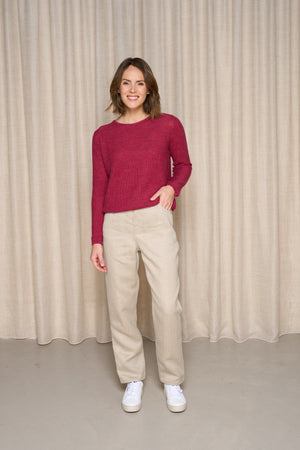 Modèle féminin présentant le jeans en lin Lutèce en Lin Naturel, coupe Mom confortable sur les hanches et étroit aux chevilles, couleur naturelle sans teinture, porté avec un pull bordeaux et des baskets blanches, souriante devant un rideau beige.