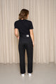 Modèle féminin de dos portant le jeans en lin Lutèce Noir coupe Mom à taille haute, couleur noir jeans, avec un t-shirt noir et des baskets blanches, sur un fond de rideau beige - Le Gaulois jeans