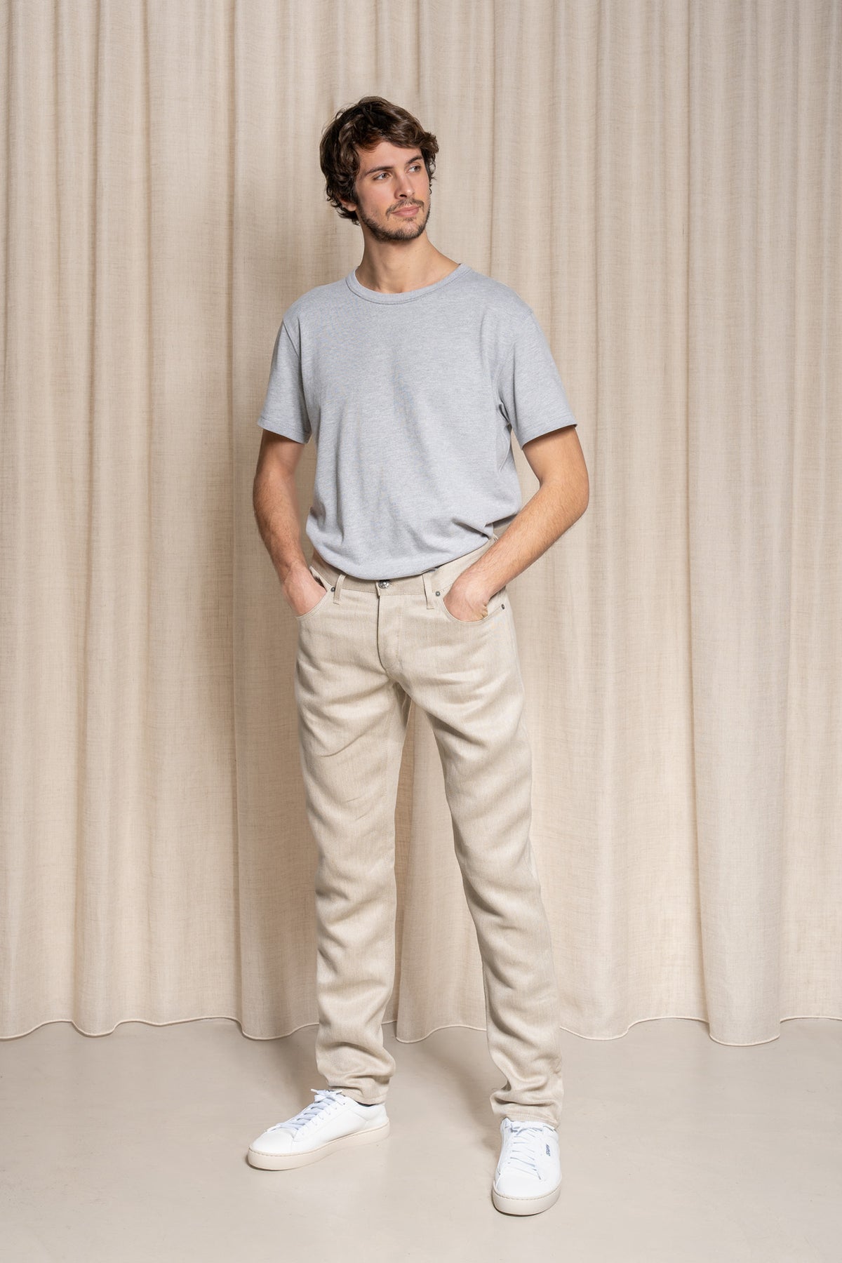 Homme debout portant le jeans en lin Massalia en Lin Naturel de couleur claire ivoire sans teinture, coupe droite légèrement effilée aux chevilles, taille mi-haute, avec un t-shirt gris et des baskets blanches, sur fond beige uni - Le Gaulois jeans