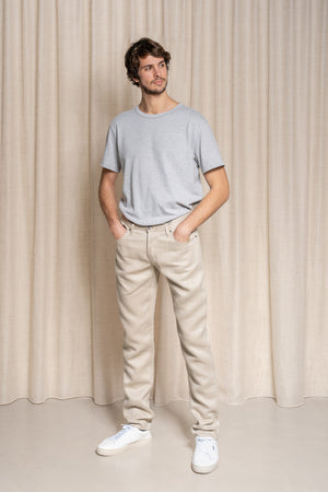 Homme debout portant le jeans en lin Massalia en Lin Naturel de couleur claire ivoire sans teinture, coupe droite légèrement effilée aux chevilles, taille mi-haute, avec un t-shirt gris et des baskets blanches, sur fond beige uni - Le Gaulois jeans
