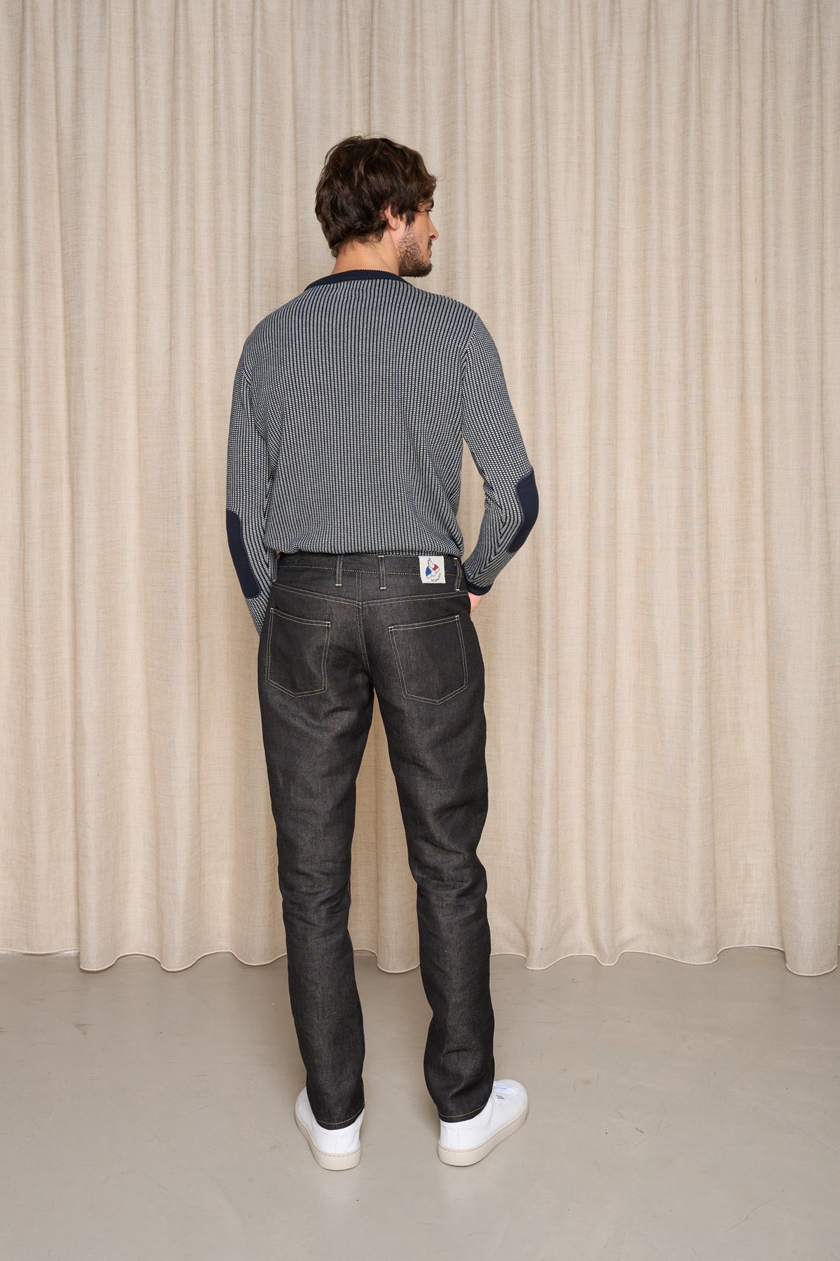 Homme de dos portant le jeans en lin modèle Massalia Noir coupe droite mi-haute, couleur noir jeans, avec un pull rayé bleu et blanc et des baskets blanches, sur fond de rideau beige et sol gris - Le Gaulois jeans