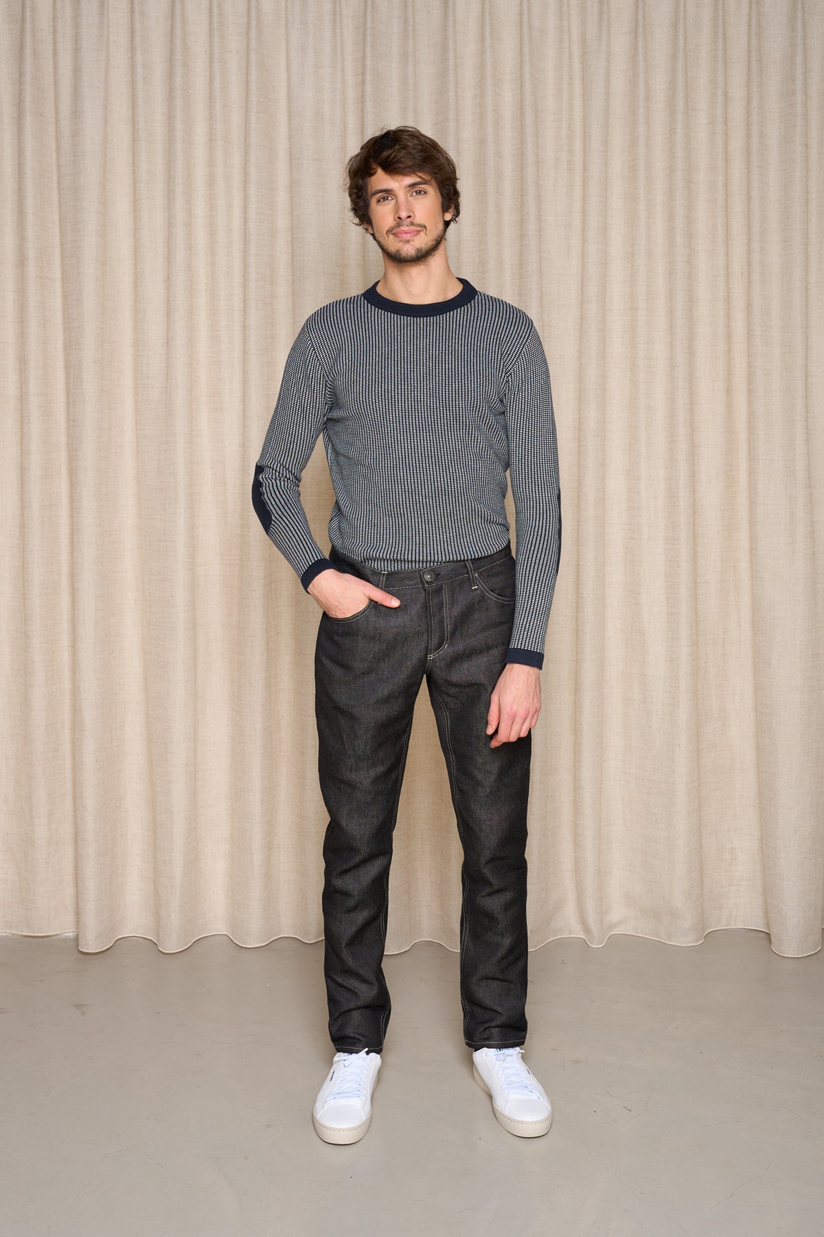 Homme présentant le jeans en lin Massalia Noir avec une coupe droite et taille mi-haute, couleur noir jeans, portant également un pull rayé et des baskets blanches, posant devant un rideau beige - Le Gaulois jeans