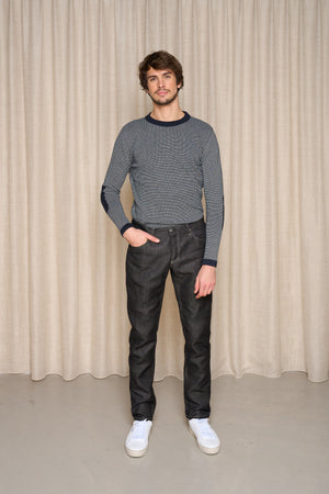 Homme présentant le jeans en lin Massalia Noir avec une coupe droite et taille mi-haute, couleur noir jeans, portant également un pull rayé et des baskets blanches, posant devant un rideau beige - Le Gaulois jeans