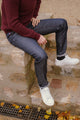 Modèle homme assis sur un rebord de pierre portant le jeans en lin et laine Narbo Bleu avec coupe droite, couleur bleu jeans, ourlet légèrement frangé aux chevilles, pull bordeaux, chaussures de sport blanches et chaussettes noires, sur fond extérieur avec feuilles d'automne au sol.