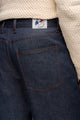 Jeans en lin et laine Narbo Bleu coupe droite avec léger effilage aux chevilles, poche arrière visible, fermeture à bouton en métal et étiquette de marque, porté sous un pull beige à grosse maille.