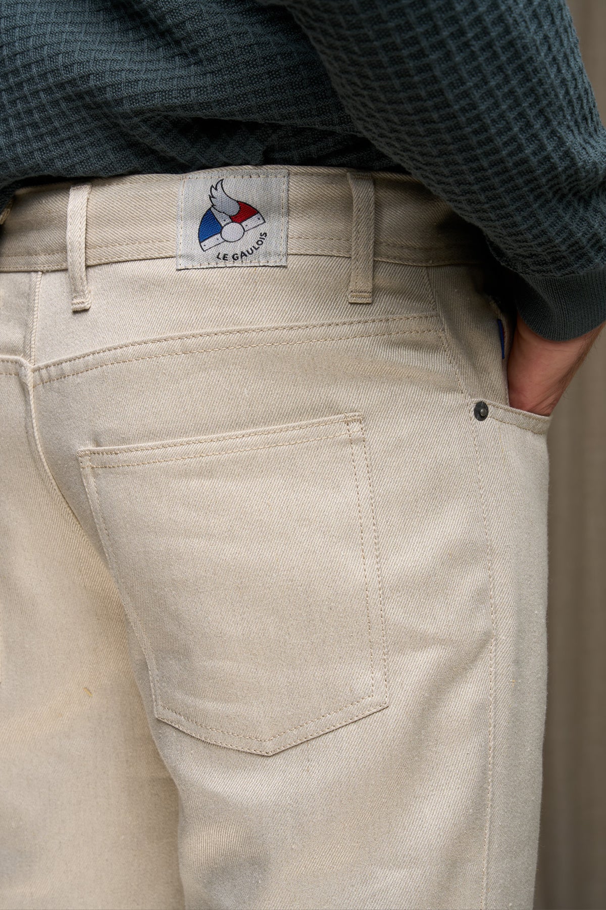 Jeans en lin et laine Narbo Naturel coupe droite avec finitions effilées aux chevilles, teinte lin naturel, bouton en métal et étiquette Le Gaulois sur la poche arrière, porté avec un haut en maille gris.