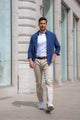 Homme marchant en ville vêtu du pantalon Divio en lin naturel de couleur ivoire, coupe près du corps, taille haute, avec chemise bleue ouverte, tee-shirt blanc, ceinture noire et baskets, reflétant l'élégance et la simplicité du jeans en lin.