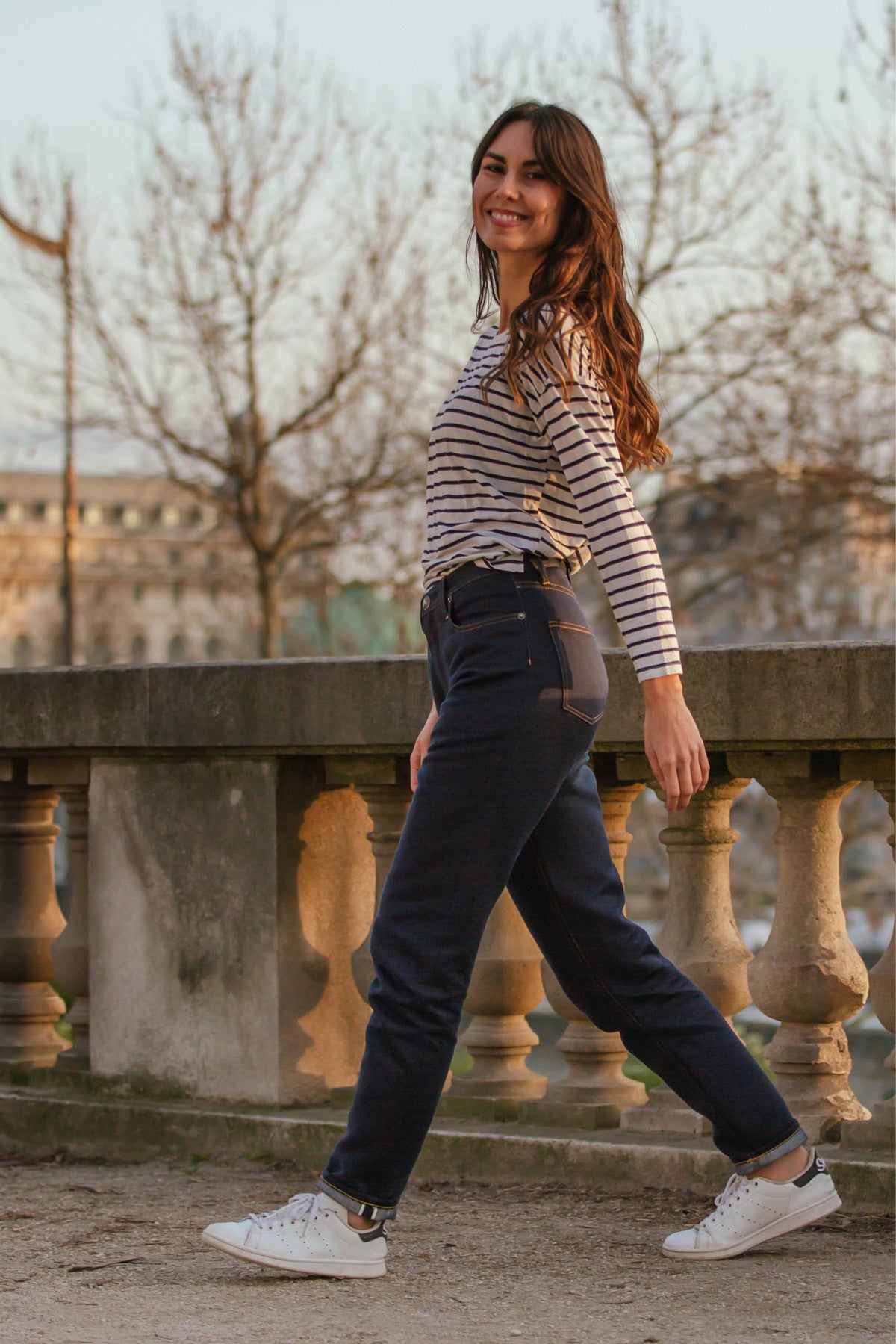 Femme souriante portant le jeans en chanvre et coton bio Isla pour femme à coupe droite, couleur bleu avec un liséré rouge sur le revers, accompagnée d'un haut rayé et de baskets blanches, en extérieur près d'une balustrade en pierre.