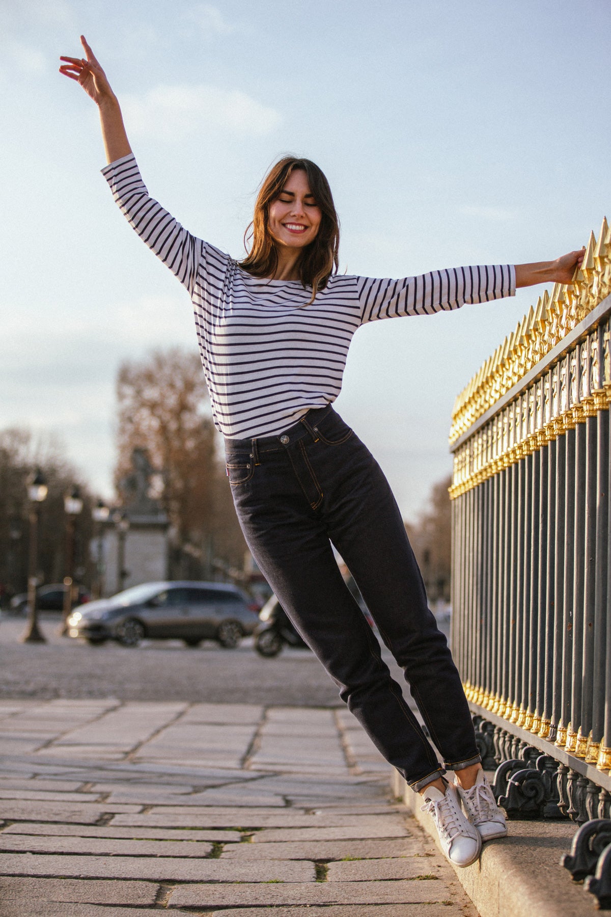 Femme souriante en mouvement, portant le jeans en chanvre et coton bio Isla pour femme à coupe droite, taille haute, avec liseré rouge visible sur le revers, accompagnée d’un haut rayé et de baskets blanches, le tout sur un trottoir avec grille dorée en arrière-plan - Le Gaulois jeans