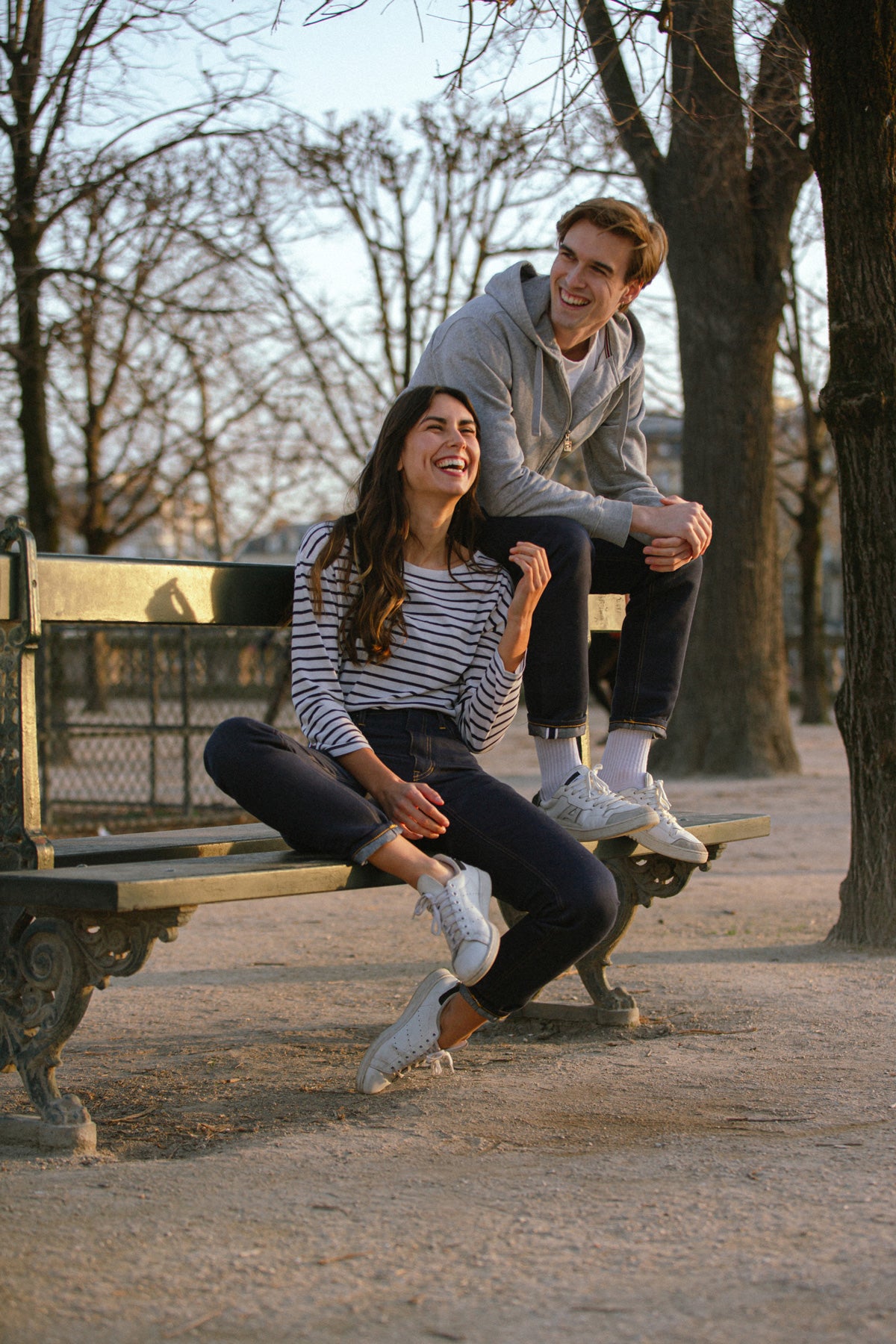 Modèle homme portant le jeans en chanvre et coton bio Isla pour homme avec coupe droite et liseré rouge, couleur bleu, accompagné d'un sweat à capuche gris et modèle femme assise riant à ses côtés, sur fond de parc avec arbres nus et banc en fer forgé