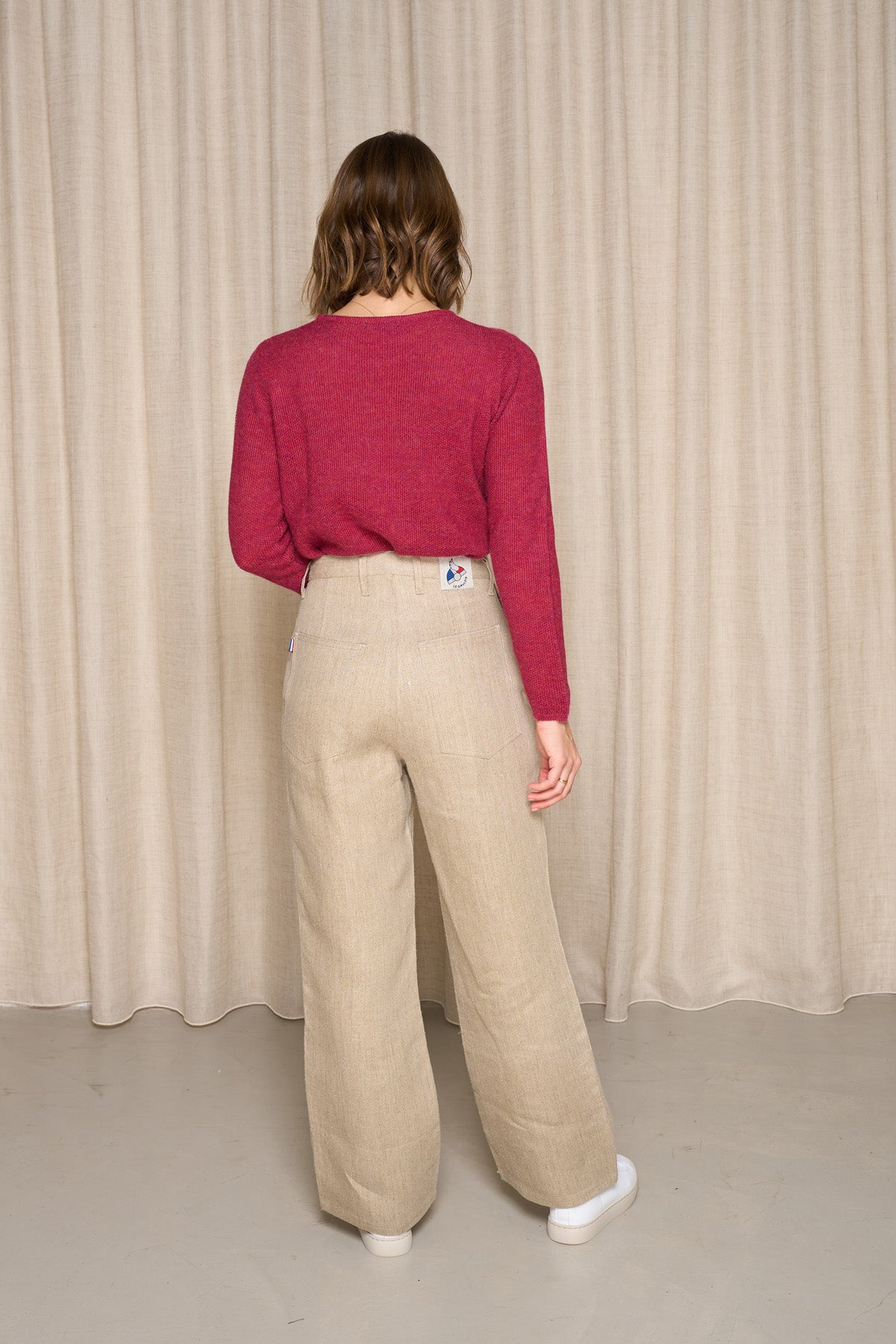 Modèle femme de dos portant le pantalon Belna en chanvre naturel avec une coupe large et taille haute, couleur naturelle sans teinture, doté d'une fermeture éclair recyclée et un bouton Diabolo en bois de buis, jeans en chanvre, sur fond de rideau beige - Le Gaulois jeans