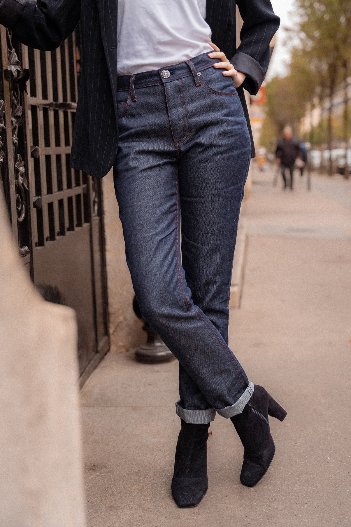 Femme debout sur un trottoir portant le jeans en lin et laine mérinos Boutae Bleu avec coupe droite et taille haute, couleur bleu jeans, fermé par glissière, associé à un tee-shirt blanc et des bottines noires, dans un cadre urbain.