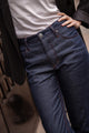 Plan rapproché d'un modèle féminin portant le jeans en lin et laine mérinos Boutae Bleu avec une coupe droite et taille haute, fermeture à glissière, couleur bleu jeans et des surpiqûres rouges, assorti à une chemise blanche et une veste noire, sur fond flou.