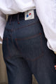 Modèle féminin portant le jeans en lin et laine mérinos Boutae Bleu avec coupe droite et taille haute, couleur bleu jeans, détail de surpiqûres contrastantes et étiquette de marque visible, accompagné d'une chemise blanche ample, sur fond neutre.