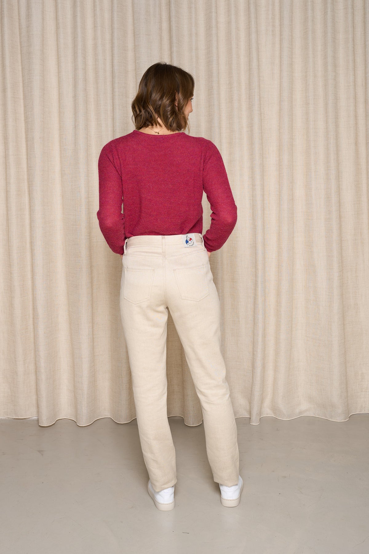 Modèle femme de dos portant le jeans en lin et laine mérinos Boutae Naturel à coupe droite et taille haute, couleur lin naturel, avec un pull bordeaux et des baskets blanches, sur fond de rideau beige - Le Gaulois jeans