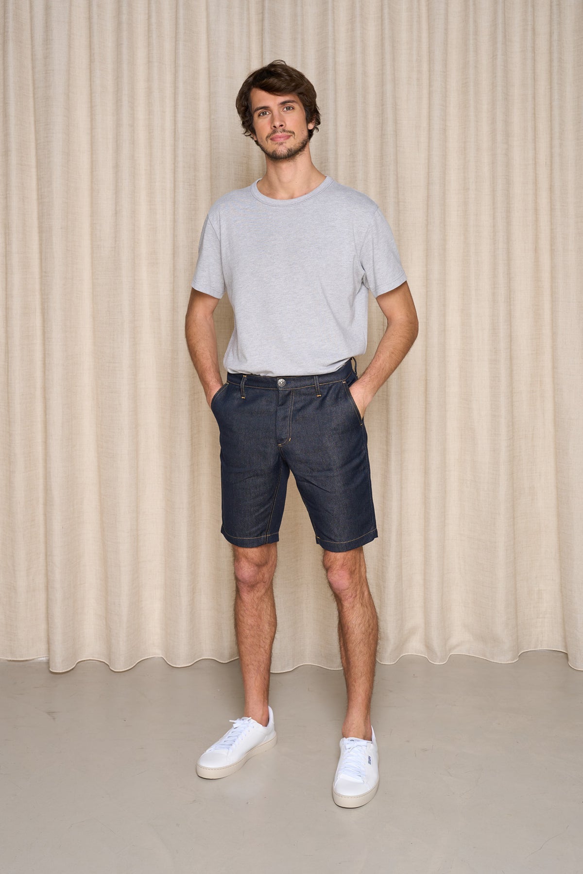 Homme portant le bermuda Cabellio en lin bleu jeans avec taille mi-haute et longueur jusqu'au genou, associé à un t-shirt gris et des baskets blanches, devant un rideau beige - Le Gaulois jeans