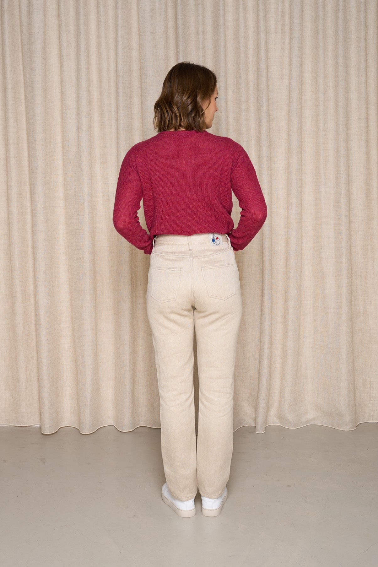 Modèle femme vue de dos portant le jeans en lin Condate en Lin Naturel à coupe droite, couleur ivoire naturelle, taille mi-haute avec fermeture à glissière, associé à un pull rouge et des baskets blanches, sur fond de rideau beige - Le Gaulois Jeans