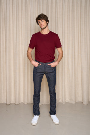 Homme debout devant un rideau de couleur beige, portant le jeans en lin et élasthanne recyclé modèle Divio bleu avec une coupe près du corps et taille haute, accompagné d'un t-shirt bordeaux et de baskets blanches - Le Gaulois jeans