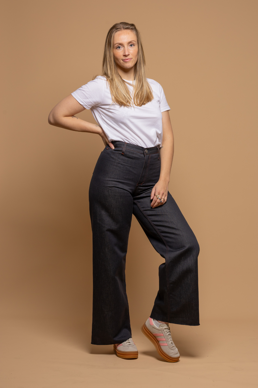 Modèle femme portant le jeans en lin et coton bio Flavia à coupe évasée en bas et taille mi-haute, de couleur bleu jeans, avec un t-shirt blanc et des baskets marrons, sur fond neutre - Le Gaulois jeans