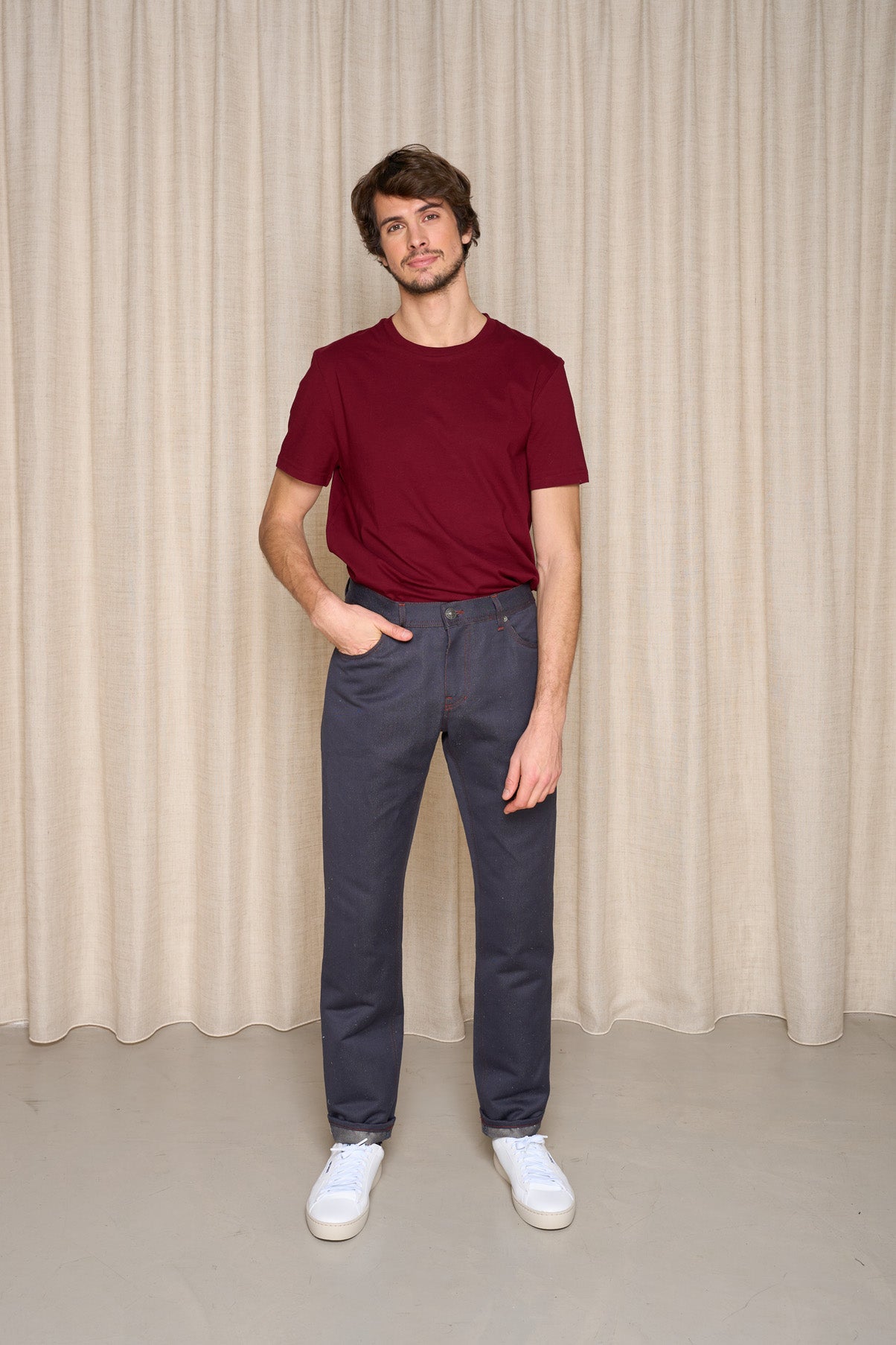 Homme présentant le jeans en chanvre et coton bio Isla pour homme couleur bleu à coupe droite, taille haute avec fermeture à glissière et liseré rouge intérieur, porté avec un t-shirt bordeaux et des baskets blanches, sur fond de rideau beige - Le Gaulois jeans