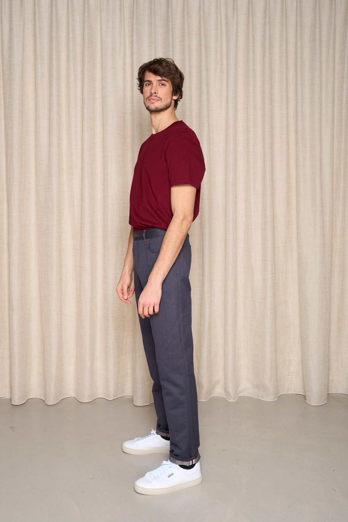 Modèle homme portant le jeans en chanvre et coton bio Isla pour homme avec coupe droite, couleur bleue et liseré rouge sur le revers, accompagné d'un t-shirt bordeaux et des baskets blanches, sur fond de rideau beige - Le Gaulois jeans
