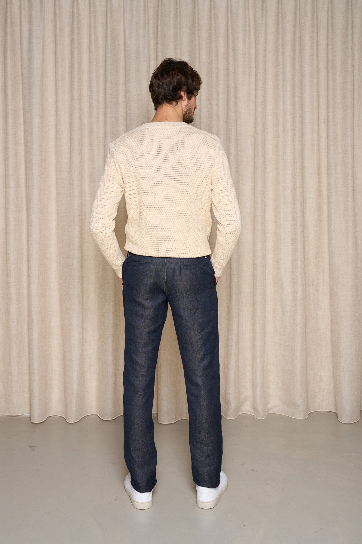 Homme de dos portant le jeans en lin Lugdunum Bleu, coupe chino, confortable aux hanches et légèrement effilé aux chevilles, taille haute, en tissu 100% lin de Normandie, couleur bleu jeans, assorti d'un pull beige et de baskets blanches, sur fond de rideau beige - Le Gaulois jeans