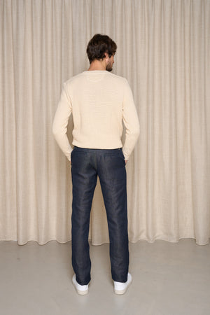 Homme de dos portant le jeans en lin Lugdunum Bleu, coupe chino, confortable aux hanches et légèrement effilé aux chevilles, taille haute, en tissu 100% lin de Normandie, couleur bleu jeans, assorti d'un pull beige et de baskets blanches, sur fond de rideau beige - Le Gaulois jeans
