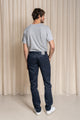 Homme de dos portant le jeans en lin Lugdunum Bleu avec coupe chino confortable, couleur bleu marine, taille haute et fermeture à glissière, associé à un t-shirt gris et des baskets blanches, sur fond de rideau beige - Le Gaulois jeans