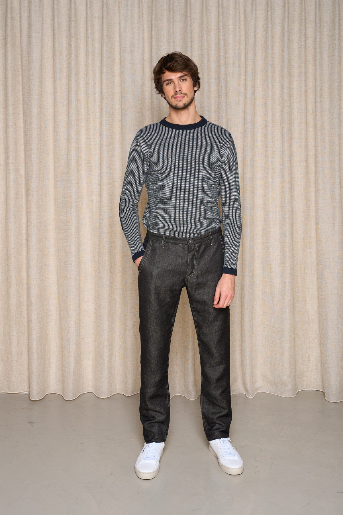 Modèle homme présentant le jeans en lin Lugdunum Noir coupe chino, confortable à la taille et légèrement effilé aux chevilles, porté avec un pull rayé et des baskets blanches, sur fond de rideau beige - Le Gaulois jeans