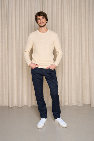 Modèle homme portant le jeans en lin Massalia Bleu à coupe droite et taille mi-haute, couleur bleu marine, accompagné d'un pull beige à mailles et des baskets blanches, sur fond de rideau beige - Le Gaulois jeans