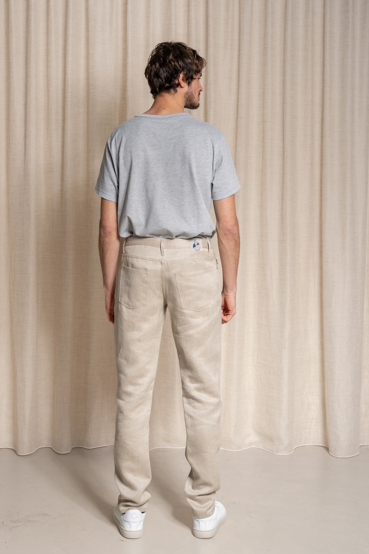 Homme de dos portant le pantalon Massalia en lin naturel beige clair ivoire, coupe droite, taille mi-haute, légèrement effilé aux chevilles, avec un t-shirt gris et des baskets blanches, sur un fond de rideau beige - Le Gaulois jeans en lin de Normandie