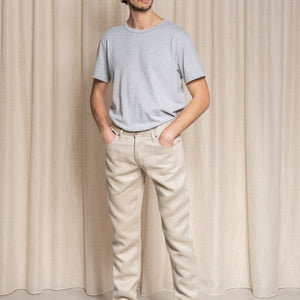 Homme debout portant le jeans en lin Massalia en Lin Naturel de couleur claire ivoire sans teinture, coupe droite légèrement effilée aux chevilles, taille mi-haute, avec un t-shirt gris et des baskets blanches, sur fond beige uni - Le Gaulois jeans