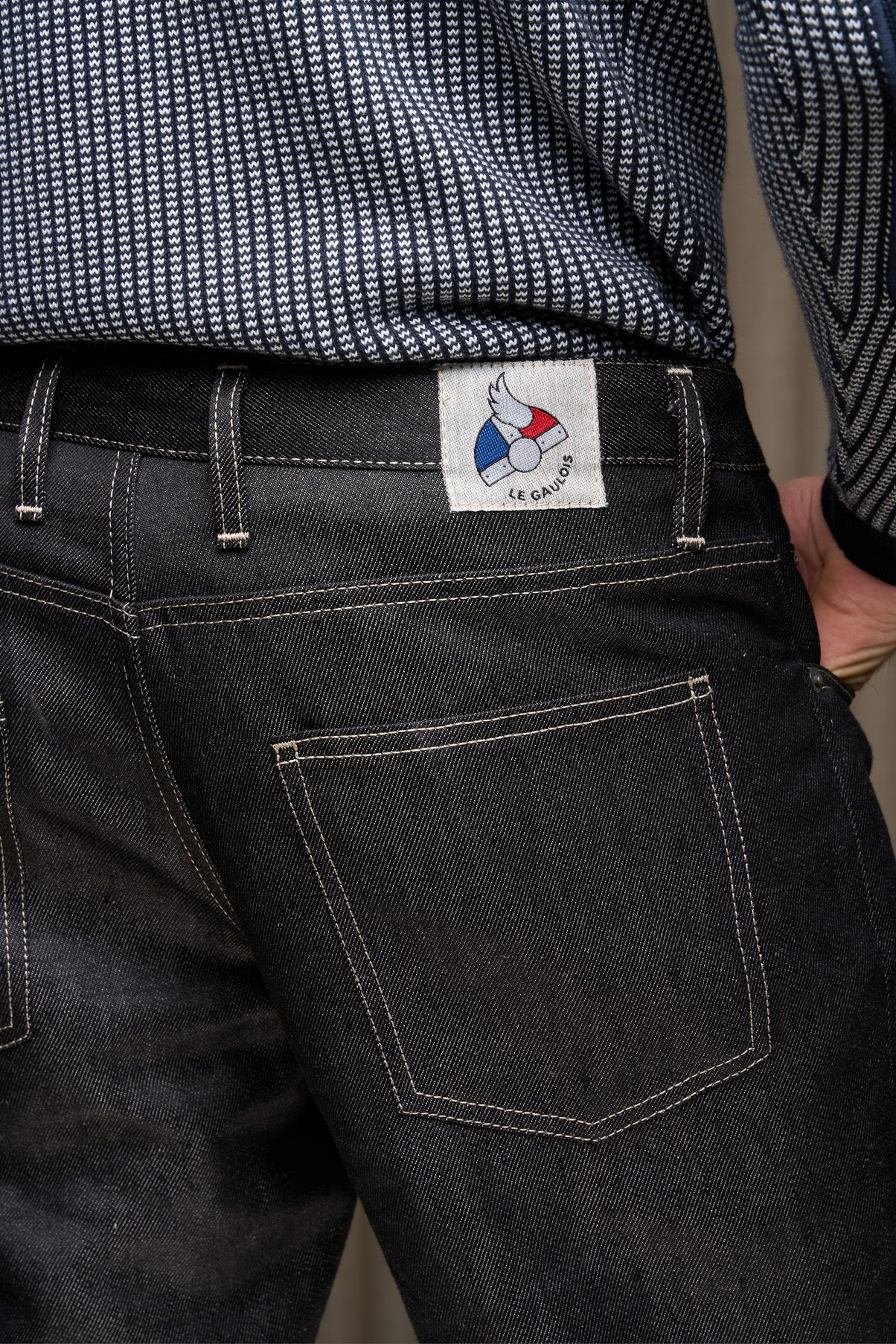 Homme de dos portant le jeans en lin Massalia Noir avec coupe droite et taille mi-haute, légèrement effilé aux chevilles, détail de la poche arrière visible et étiquette Le Gaulois jeans, porté sous une chemise à motif chevron sur fond gris.