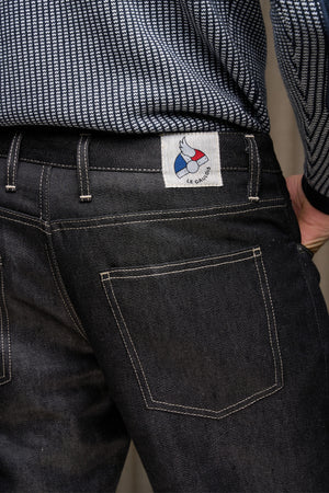 Homme de dos portant le jeans en lin Massalia Noir avec coupe droite et taille mi-haute, légèrement effilé aux chevilles, détail de la poche arrière visible et étiquette Le Gaulois jeans, porté sous une chemise à motif chevron sur fond gris.