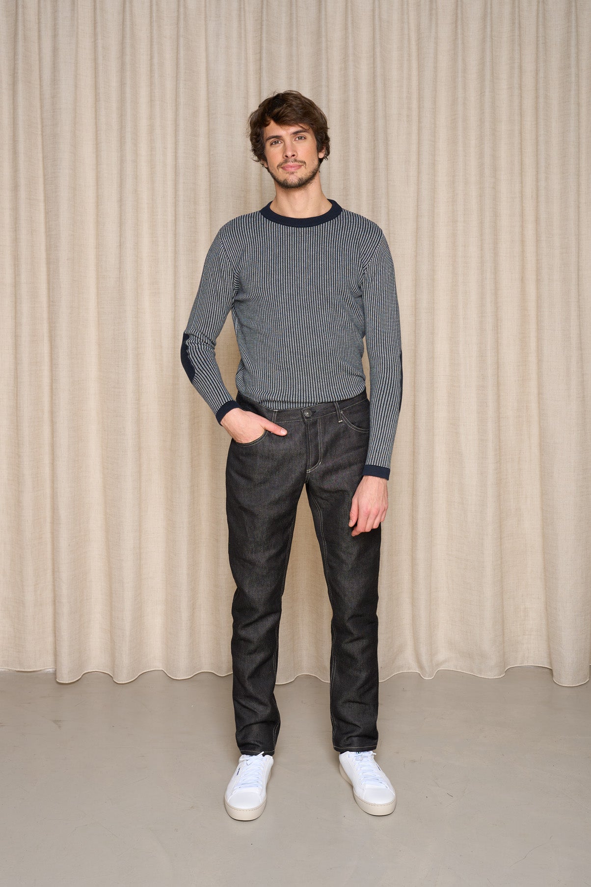 Homme présentant le jeans en lin Massalia Noir avec une coupe droite et taille mi-haute, couleur noir jeans, portant également un pull rayé et des baskets blanches, posant devant un rideau beige - Le Gaulois jeans
