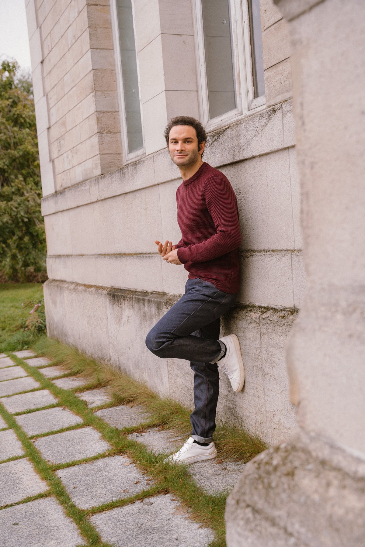 Homme appuyé contre un mur en pierre portant le jeans en lin et laine mérinos Narbo Bleu à coupe droite, couleur bleu jeans, avec un pull bordeaux et des baskets blanches, dans un cadre extérieur avec des pavés et de l'herbe.
