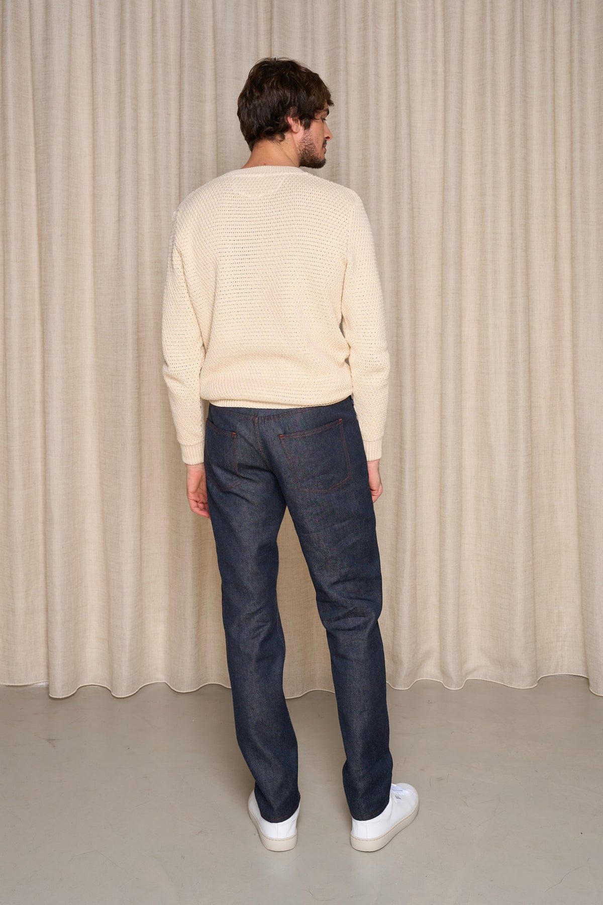 Homme de dos portant le jeans en lin et laine mérinos Narbo Bleu à coupe droite avec léger effilage aux chevilles, couleur bleu jeans, complété par un pull beige et des baskets blanches, sur fond de rideau beige - Le Gaulois jeans