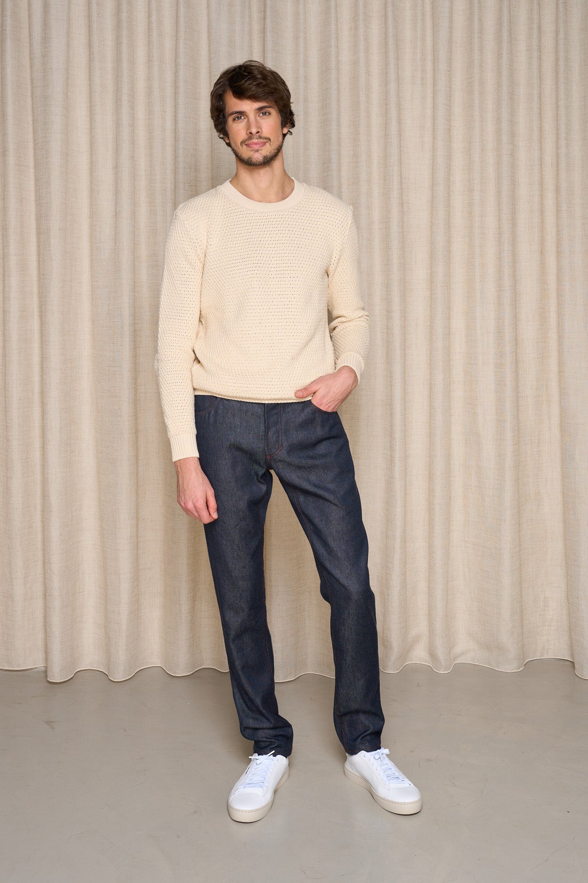 Modèle homme présentant le pantalon Narbo Bleu, jeans en Lin de Normandie et Laine Mérinos française, coupe droite avec effilage léger aux chevilles, fermeture à bouton métallique, couleur bleu jeans, porté avec un pull beige et des baskets blanches, sur un fond de rideau beige - Le Gaulois jeans.