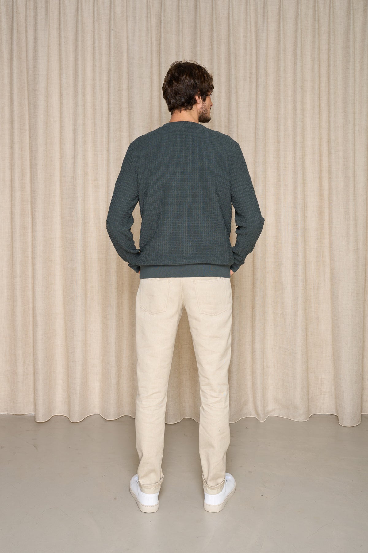 Homme de dos portant un jeans en lin et laine mérinos Narbo Naturel à coupe droite, couleur lin naturel, avec finition effilée aux chevilles, un pull bleu foncé et des baskets blanches, sur fond de rideau beige - Le Gaulois jeans