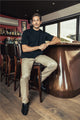 Homme portant le jeans en chanvre L'Aurelien de couleur naturelle avec coupe droite, poches italiennes, bouton en bois de buis et fermeture éclair recyclée, assis sur un tabouret dans un bar chic.