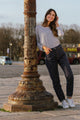 Modèle féminin posant avec le jeans en chanvre et coton bio Isla pour femme, de couleur bleue, coupe droite et taille haute avec fermeture à glissière et liseré rouge sur le revers, porté avec un haut à rayures blanches et bleues, debout près d'une colonne décorée sur un trottoir parisien.