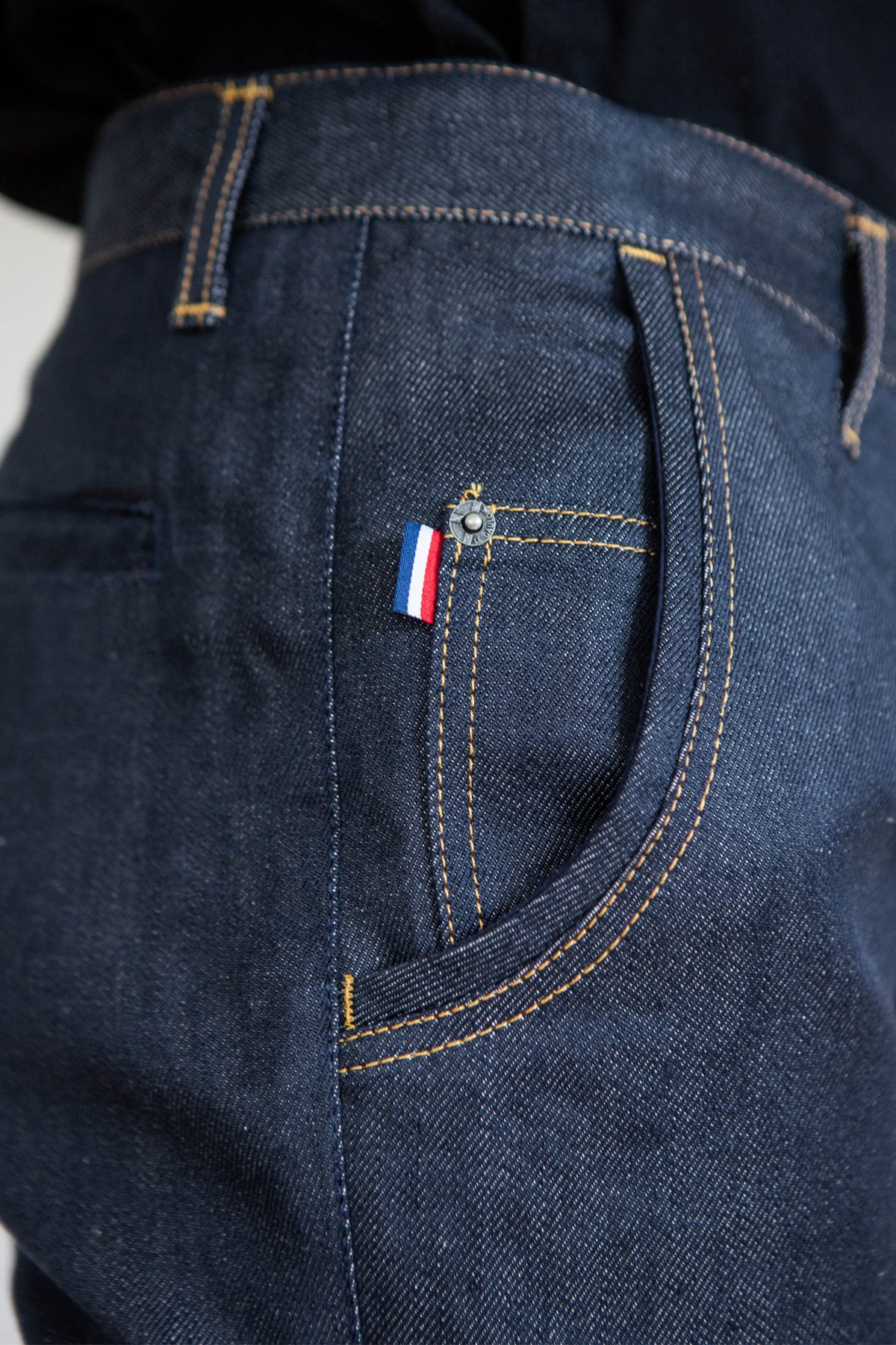 Modèle homme portant le jeans en lin Lugdunum Bleu avec coupe chino, détail d'un écusson tricolore et coutures contrastantes, fabrications française et tissu de Normandie, sur un fond uni.